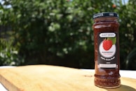 A jar of raspberry fruit spread labeled 'Blast Berry' is placed on a wooden surface. The background consists of blurred green foliage, indicating an outdoor setting.