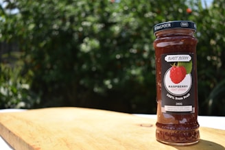 A jar of raspberry fruit spread labeled 'Blast Berry' is placed on a wooden surface. The background consists of blurred green foliage, indicating an outdoor setting.