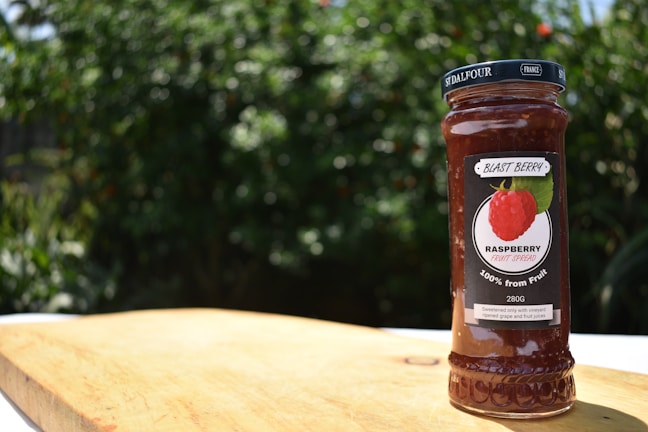 A jar of raspberry fruit spread labeled 'Blast Berry' is placed on a wooden surface. The background consists of blurred green foliage, indicating an outdoor setting.