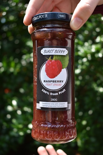 A hand holds a glass jar of raspberry fruit spread labeled 'Blast Berry'. The jar is topped with a black lid featuring branding indicating it is from 'France'. The label prominently displays a raspberry image and notes '100% from Fruit' and '280G'. The background is a blurred green foliage, suggesting an outdoor setting.