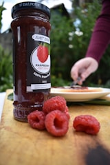 A jar of raspberry fruit spread sits on a wooden surface with fresh raspberries scattered in front. A person in the background, wearing a maroon sleeve, reaches toward a plate with food, possibly using a utensil.