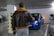 A person wearing a brown leather jacket with a shearling collar is standing in a parking garage. They are facing a blue Fiat car with its headlights on, parked between other vehicles. The setting is an indoor parking lot with concrete floors and a white pillar marked with black and yellow caution stripes.