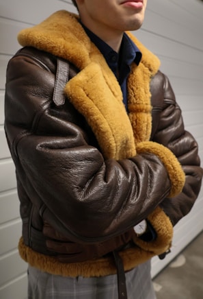 A person wearing a brown leather jacket with fur lining. The jacket has a classic aviator style and appears warm and cozy. The person stands with arms crossed, conveying a relaxed or confident demeanor.