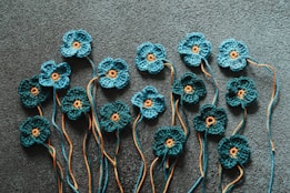 A series of intricately crocheted blue flowers with orange centers are displayed against a textured dark background. Each flower is attached to strands of yarn that extend in various directions, adding a layered and artistic quality to the arrangement.