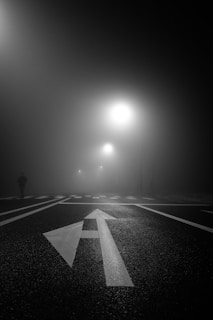 A dark, moody cinematic scene showing a shadowy figure standing at a foggy crossroads at night.