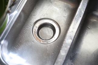 A stainless steel kitchen sink with droplets of water scattered across its surface. The metal drain is prominently visible in the center, with a circular perforated cover.