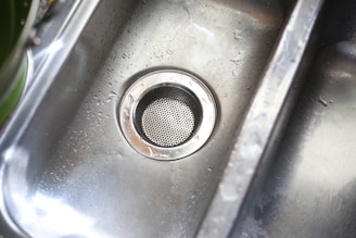 A stainless steel kitchen sink with droplets of water scattered across its surface. The metal drain is prominently visible in the center, with a circular perforated cover.