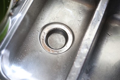 A stainless steel kitchen sink with droplets of water scattered across its surface. The metal drain is prominently visible in the center, with a circular perforated cover.