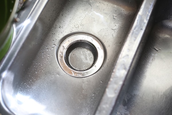 A stainless steel kitchen sink with droplets of water scattered across its surface. The metal drain is prominently visible in the center, with a circular perforated cover.