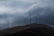 Large wind turbines are standing on rolling hills under a cloudy sky. The landscape is expansive, with the turbines evenly spaced across the terrain, suggesting a focus on renewable energy.