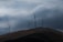 Large wind turbines are standing on rolling hills under a cloudy sky. The landscape is expansive, with the turbines evenly spaced across the terrain, suggesting a focus on renewable energy.