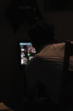 A person is sitting in a dimly lit room, facing a computer screen that displays various icons and a blurred image. The light from the screen partially illuminates the person's back and the chair they are sitting on.