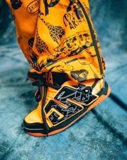 A close-up view of a vibrant orange and black sneaker with intricate patterns and graffiti-style designs. The shoe features bold shapes and lines, with visible text and artwork printed on the upper part. The background is a textured blue fabric.
