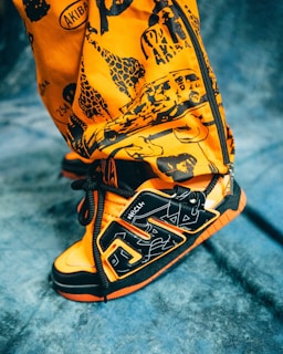 A close-up view of a vibrant orange and black sneaker with intricate patterns and graffiti-style designs. The shoe features bold shapes and lines, with visible text and artwork printed on the upper part. The background is a textured blue fabric.