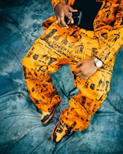 A person is wearing a bright orange outfit with black graphic designs featuring various silhouettes and shapes, including buildings and giraffes. The outfit includes a jacket and pants, and the person holds a black device in one hand. A silver watch adorns the wrist, and the feet are wearing matching orange and black sneakers. The background is a textured blue fabric.