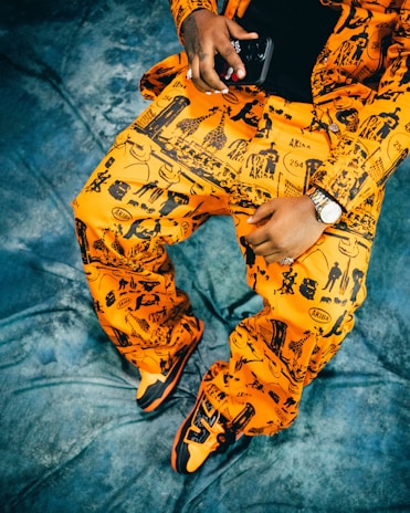 A person is wearing a bright orange outfit with black graphic designs featuring various silhouettes and shapes, including buildings and giraffes. The outfit includes a jacket and pants, and the person holds a black device in one hand. A silver watch adorns the wrist, and the feet are wearing matching orange and black sneakers. The background is a textured blue fabric.