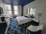 A modern hotel room with a sleek, minimalist design featuring a large bed with white linens and a navy bolster pillow. The room has a patterned blue carpet and a navy blue ceiling that contrasts with the white walls. There is a large mirror above the bed and two bedside tables with lamps. Large windows with cityscape views provide natural light. Additional furnishings include a white chair with navy accents and a work desk with a television and a coffee machine nearby. Exposed brick walls add a touch of industrial style.