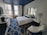A modern hotel room with a sleek, minimalist design featuring a large bed with white linens and a navy bolster pillow. The room has a patterned blue carpet and a navy blue ceiling that contrasts with the white walls. There is a large mirror above the bed and two bedside tables with lamps. Large windows with cityscape views provide natural light. Additional furnishings include a white chair with navy accents and a work desk with a television and a coffee machine nearby. Exposed brick walls add a touch of industrial style.
