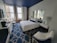 A modern hotel room with a sleek, minimalist design featuring a large bed with white linens and a navy bolster pillow. The room has a patterned blue carpet and a navy blue ceiling that contrasts with the white walls. There is a large mirror above the bed and two bedside tables with lamps. Large windows with cityscape views provide natural light. Additional furnishings include a white chair with navy accents and a work desk with a television and a coffee machine nearby. Exposed brick walls add a touch of industrial style.