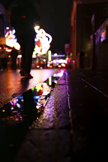 Nighttime cityscape with neon signs reflecting off wet pavement, a silhouette wearing a Nomadika tee walking confidently.