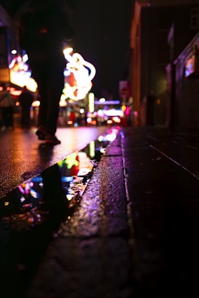Urban street scene at night with neon signs reflecting on wet pavement.