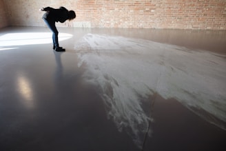 A person is bending over to examine a pattern or texture on a smooth, reflective floor. The space appears to be minimalistic with a brick wall in the background. Light is casting through a window, creating shadows and highlights on the floor.