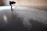 A person is bending over to examine a pattern or texture on a smooth, reflective floor. The space appears to be minimalistic with a brick wall in the background. Light is casting through a window, creating shadows and highlights on the floor.