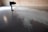 A person is bending over to examine a pattern or texture on a smooth, reflective floor. The space appears to be minimalistic with a brick wall in the background. Light is casting through a window, creating shadows and highlights on the floor.