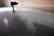 A person is bending over to examine a pattern or texture on a smooth, reflective floor. The space appears to be minimalistic with a brick wall in the background. Light is casting through a window, creating shadows and highlights on the floor.
