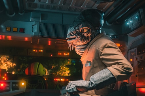 A humanoid creature with an aquatic-like head and large eyes is sitting inside a futuristic control room. The environment is dimly lit with various colorful buttons and indicators glowing on the control panel. Outside the window, some illuminated shapes or structures can be seen.