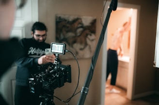 A film set with a camera on a tripod is positioned in the foreground. A person wearing glasses is looking at a smartphone. Another person is visible in the background, possibly in a bathroom, blurred and partially out of focus. The setting appears to be indoors with a painting of horses on the wall.