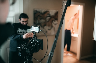 A film set with a camera on a tripod is positioned in the foreground. A person wearing glasses is looking at a smartphone. Another person is visible in the background, possibly in a bathroom, blurred and partially out of focus. The setting appears to be indoors with a painting of horses on the wall.