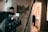 A film set with a camera on a tripod is positioned in the foreground. A person wearing glasses is looking at a smartphone. Another person is visible in the background, possibly in a bathroom, blurred and partially out of focus. The setting appears to be indoors with a painting of horses on the wall.