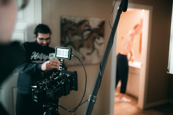 A film set with a camera on a tripod is positioned in the foreground. A person wearing glasses is looking at a smartphone. Another person is visible in the background, possibly in a bathroom, blurred and partially out of focus. The setting appears to be indoors with a painting of horses on the wall.