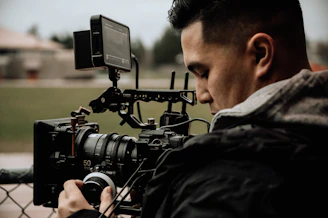 A person is operating a professional video camera, focusing intently on the task. The setting appears to be outdoors, as suggested by the blurred background of greenery and buildings. The camera features several components, including a large lens, dials, and a monitor mounted on top.