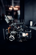 A professional video camera setup is positioned on a dark surface. The camera has multiple lenses and attachments, including a monitor screen displaying an image. The area around the camera is dimly lit, with some blurred background elements and light sources visible.