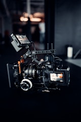 A professional video camera setup is positioned on a dark surface. The camera has multiple lenses and attachments, including a monitor screen displaying an image. The area around the camera is dimly lit, with some blurred background elements and light sources visible.