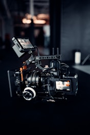 A professional video camera setup is positioned on a dark surface. The camera has multiple lenses and attachments, including a monitor screen displaying an image. The area around the camera is dimly lit, with some blurred background elements and light sources visible.
