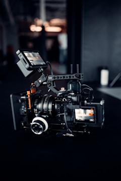 A professional video camera setup is positioned on a dark surface. The camera has multiple lenses and attachments, including a monitor screen displaying an image. The area around the camera is dimly lit, with some blurred background elements and light sources visible.