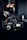 A professional video camera setup is positioned on a dark surface. The camera has multiple lenses and attachments, including a monitor screen displaying an image. The area around the camera is dimly lit, with some blurred background elements and light sources visible.
