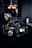 A professional video camera setup is positioned on a dark surface. The camera has multiple lenses and attachments, including a monitor screen displaying an image. The area around the camera is dimly lit, with some blurred background elements and light sources visible.