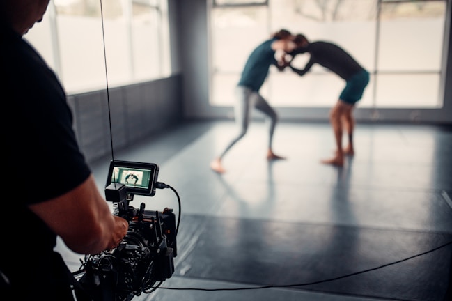 A photographer or videographer is capturing footage of two individuals engaged in a grappling or wrestling practice session on a mat in a spacious room with large windows. The camera equipment is prominently in the foreground, focusing on the action in the background, which appears slightly blurred.