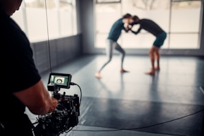 A photographer or videographer is capturing footage of two individuals engaged in a grappling or wrestling practice session on a mat in a spacious room with large windows. The camera equipment is prominently in the foreground, focusing on the action in the background, which appears slightly blurred.