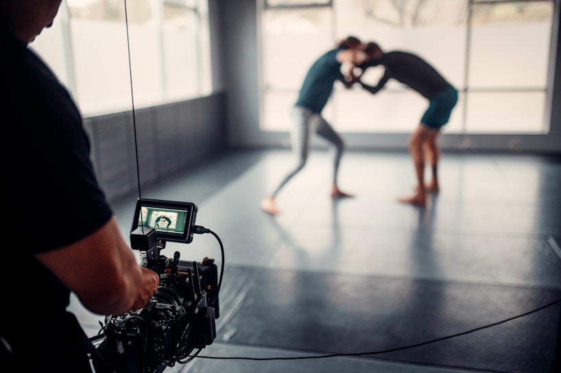 A photographer or videographer is capturing footage of two individuals engaged in a grappling or wrestling practice session on a mat in a spacious room with large windows. The camera equipment is prominently in the foreground, focusing on the action in the background, which appears slightly blurred.