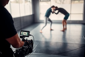 A photographer or videographer is capturing footage of two individuals engaged in a grappling or wrestling practice session on a mat in a spacious room with large windows. The camera equipment is prominently in the foreground, focusing on the action in the background, which appears slightly blurred.
