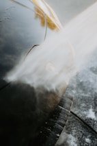 A close-up view of a car being washed with a stream of water directed at the front. The water creates a misty effect as it hits the car's surface, which is glossy and dark in color. The image highlights the car's headlight and the smooth contours of its body.