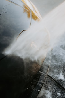 A close-up view of a car being washed with a stream of water directed at the front. The water creates a misty effect as it hits the car's surface, which is glossy and dark in color. The image highlights the car's headlight and the smooth contours of its body.
