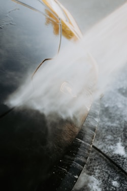 A close-up view of a car being washed with a stream of water directed at the front. The water creates a misty effect as it hits the car's surface, which is glossy and dark in color. The image highlights the car's headlight and the smooth contours of its body.