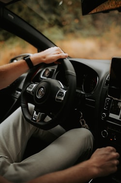 A person is sitting in the driver's seat of a car, holding the steering wheel with one hand. The interior of the vehicle includes a dashboard with visible controls and a mounted screen. The surroundings outside the window appear to be blurred, indicating motion or focus on the interior.