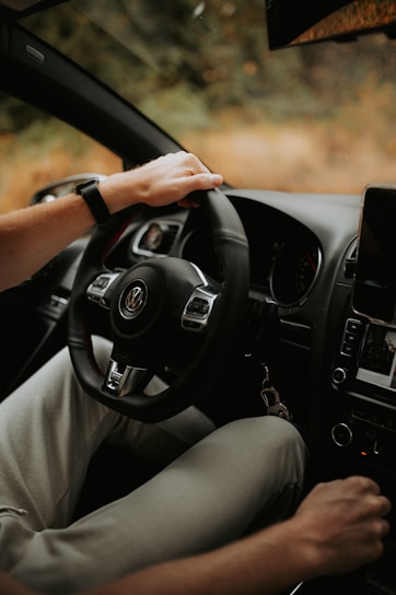 A person is sitting in the driver's seat of a car, holding the steering wheel with one hand. The interior of the vehicle includes a dashboard with visible controls and a mounted screen. The surroundings outside the window appear to be blurred, indicating motion or focus on the interior.